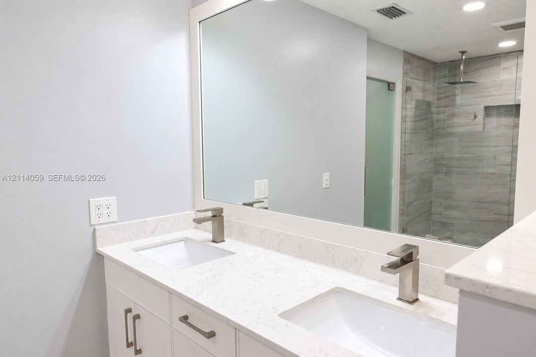 Double vanity featuring white cabinetry, stone-finish countertops, rectangular undermount sinks, and brushed-finish faucets, paired with a walk-in shower encased in glass with tile wall surfacing and a built-in recessed shelf