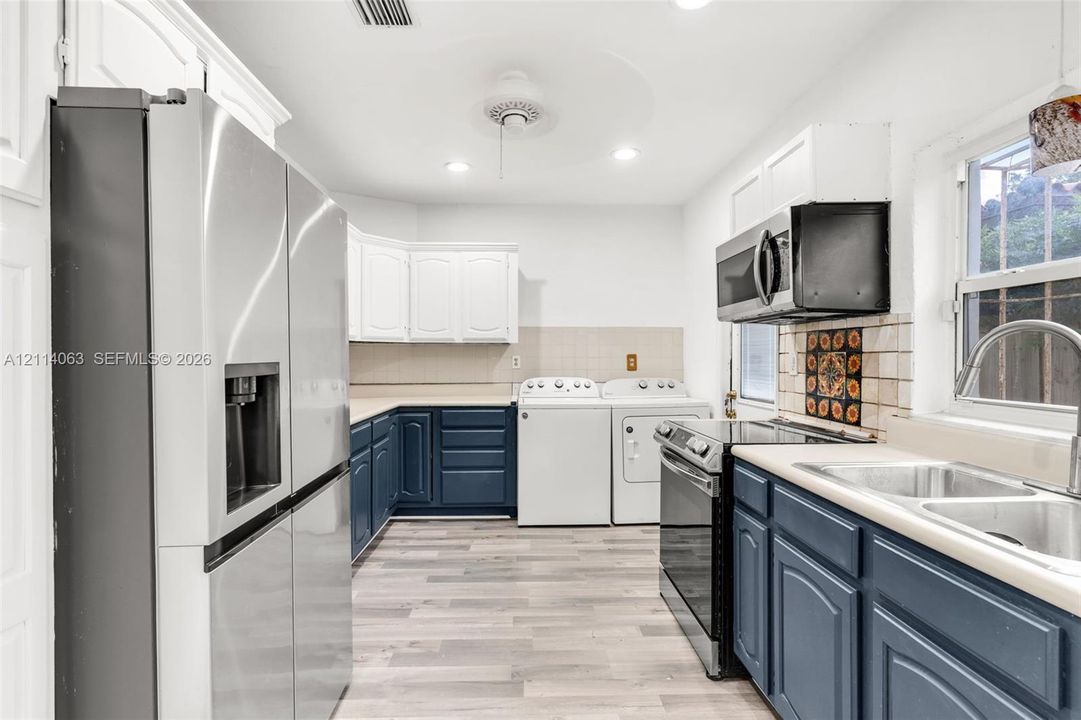 Kitchen featuring stainless steel appliances, blue and white cabinetry, wood-finish flooring, double-basin sink, and integrated laundry area