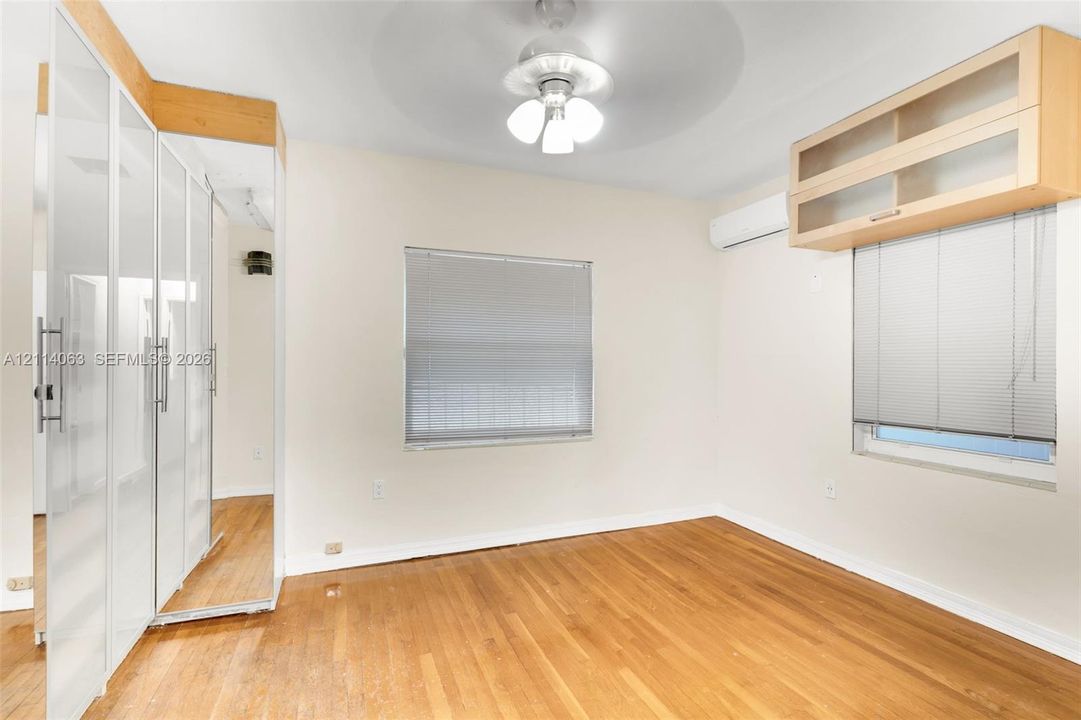 Room featuring wood-finish flooring, built-in floor-to-ceiling wardrobe cabinetry with mirrored doors, wall-mounted ductless climate control unit, and overhead ceiling fan with light fixture
