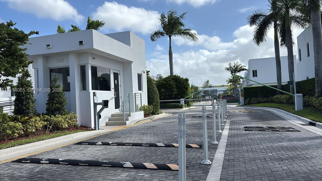 Gated entry featuring a controlled-access security booth, paved driveway, barrier arm, and tropical landscaping