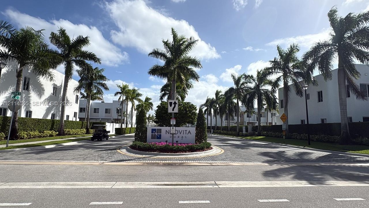 Gated community entrance featuring a paved roundabout, professional landscaping, mature palm trees, and manicured hedges