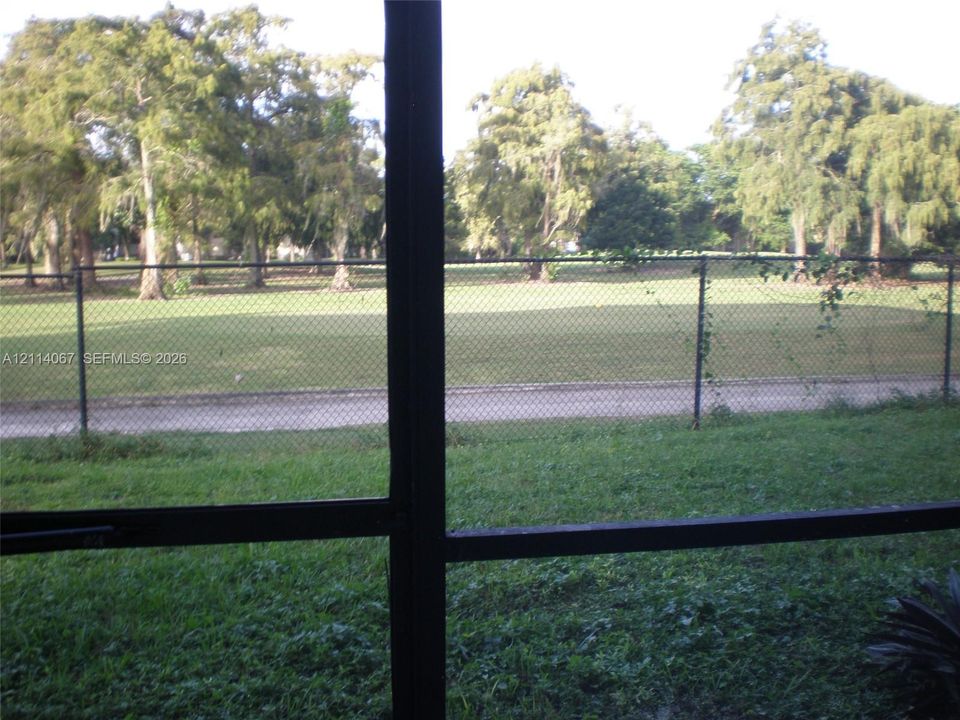 Screened enclosure overlooking a grass lawn, paved pathway, chain-link perimeter fencing, and mature trees