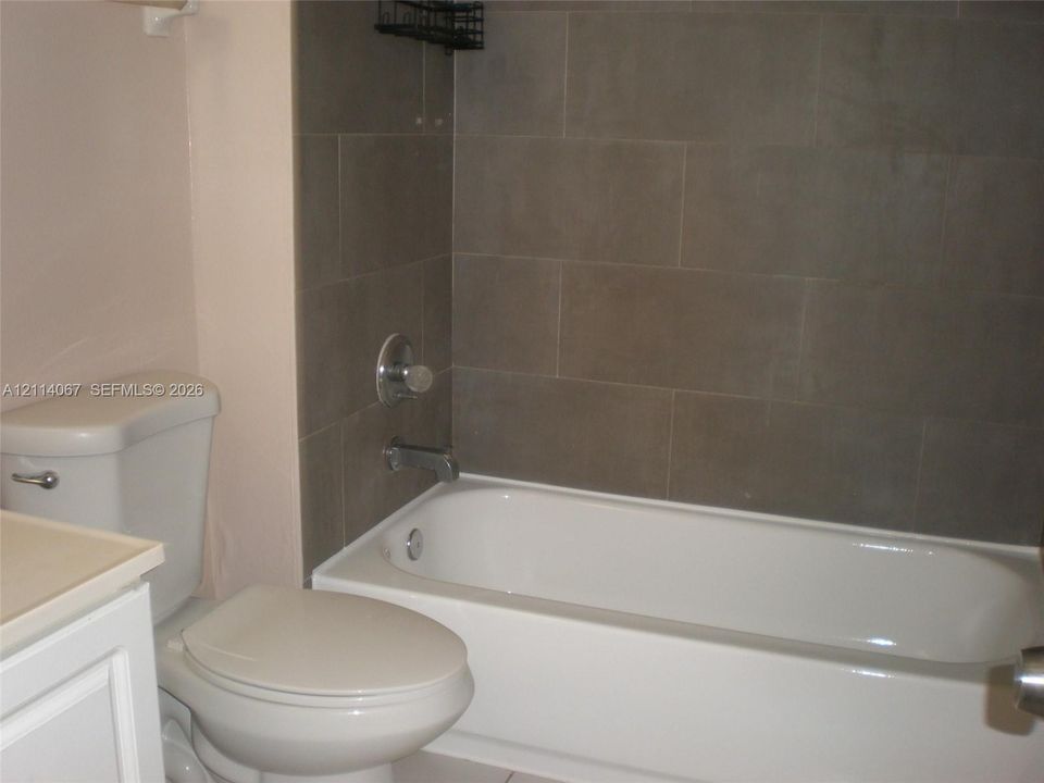 White vanity cabinet, standard porcelain toilet, and bathtub with large-format gray tile surround and integrated metal fixtures