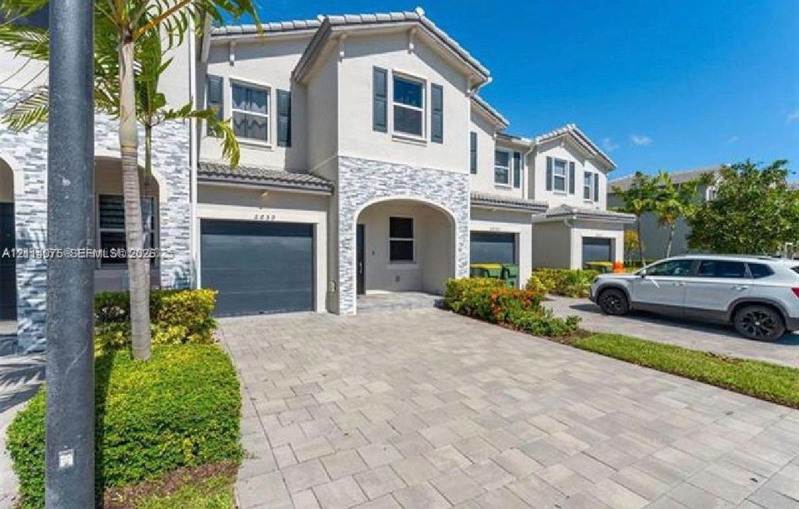 Two-story exterior featuring light-colored stucco finish, stone veneer accents, paver driveway, attached garage, and tiled roof