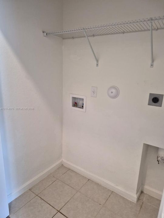 Utility space featuring ceramic tile flooring, a recessed water supply box, an electrical outlet, a dedicated power connection, and a mounted wire shelving unit