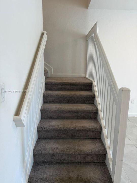 Staircase featuring neutral-toned carpeting, a white wooden handrail, and a vertical baluster railing