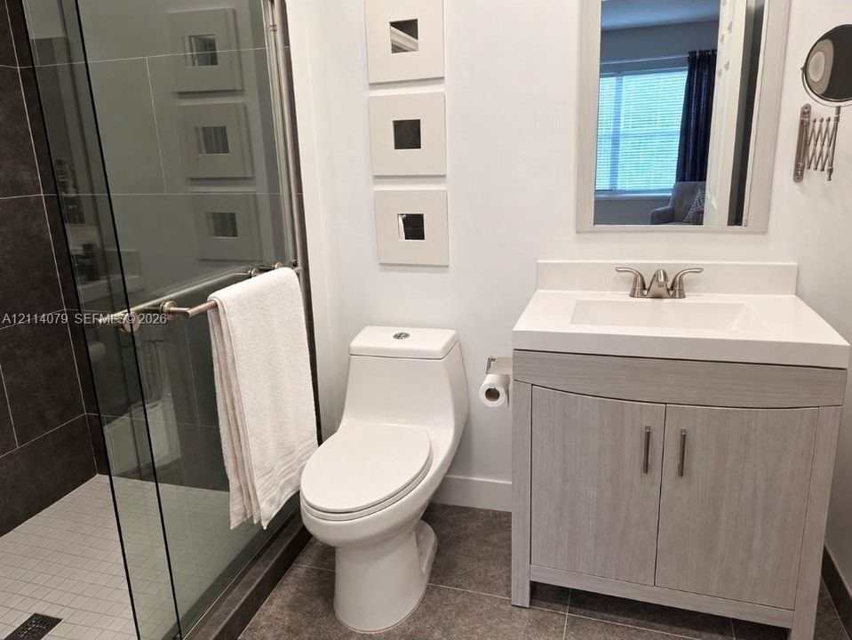 Bathroom featuring a wood-finish vanity with a rectangular sink, glass-enclosed shower with dark tile surround, and neutral floor tile