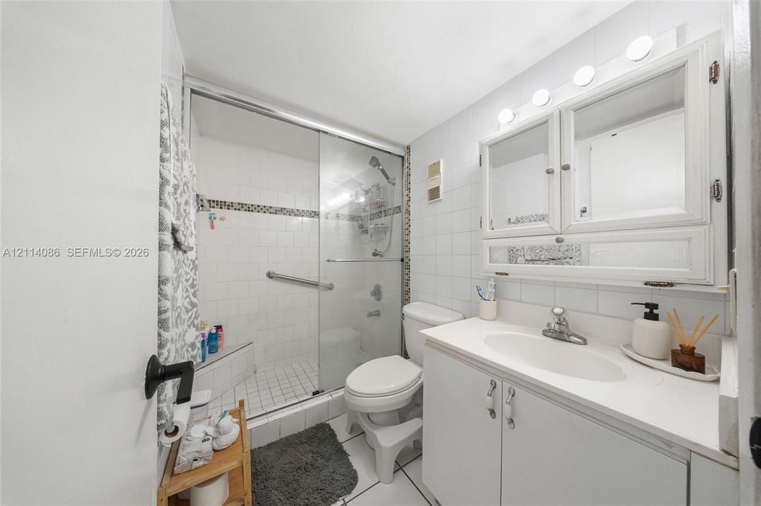 Bathroom featuring a vanity with white cabinetry, a surface-mounted medicine cabinet, a tiled shower enclosure with a glass door, and wall-mounted light fixtures