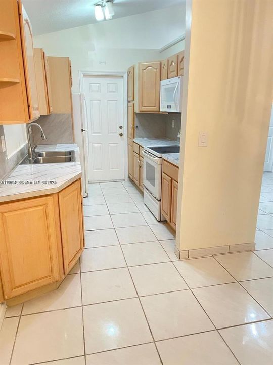 Kitchen featuring light wood cabinetry, stone-finish countertops, stainless steel dual-basin sink with arched faucet, ceramic tile flooring, and integrated white appliances