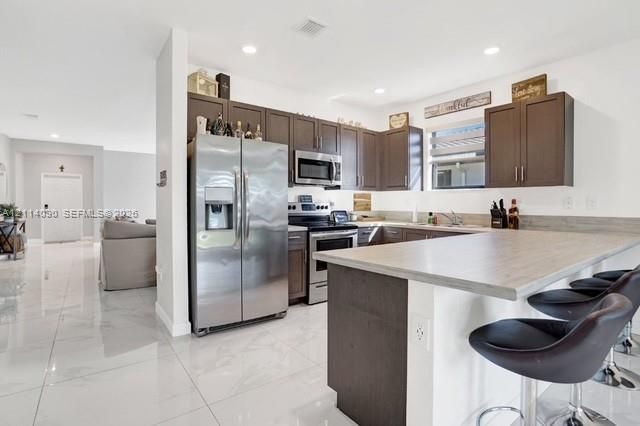 Kitchen featuring dark-finish cabinetry, stainless steel appliances, a peninsula with a stone-finish countertop, and light-toned tile flooring