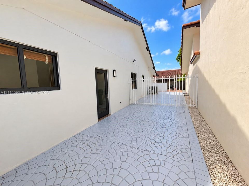 Side exterior featuring decorative stamped concrete paving, white metal perimeter fencing, wall-mounted light fixtures, and a glass-paneled entry door