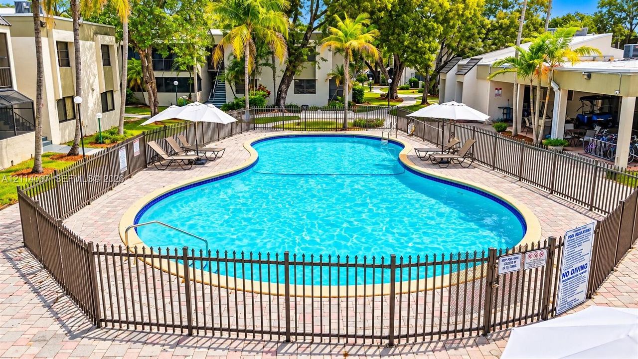 Community swimming pool featuring a paved deck, perimeter metal fencing, and surrounding mature palm trees