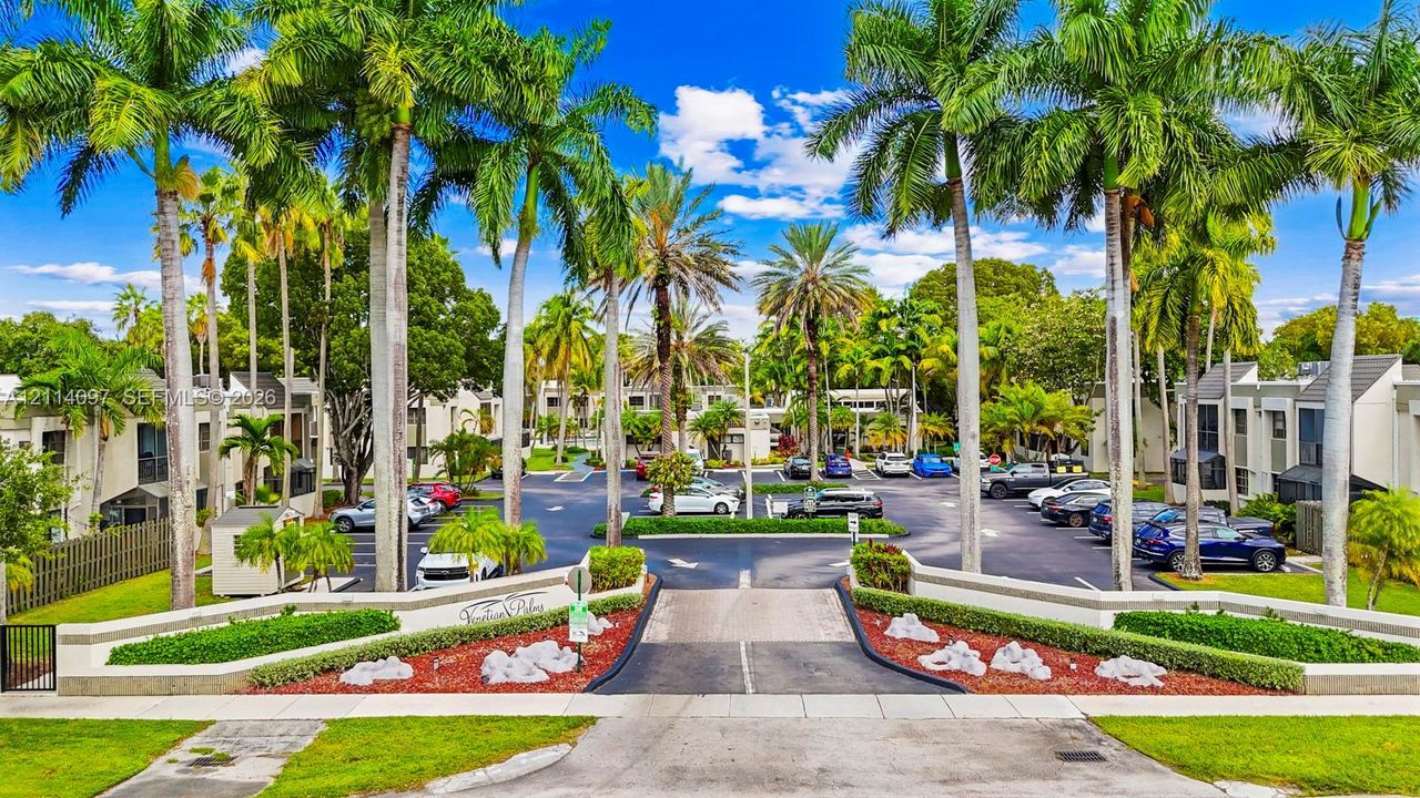 Gated community entrance featuring masonry pillars, manicured boxwood hedges, mature palm trees, and asphalt driveway