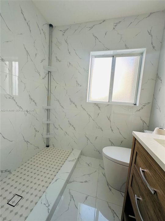 Walk-in shower featuring floor-to-ceiling large-format tile with grey veining, a geometric mosaic tile shower floor, a light-toned wood-finish vanity with a solid-surface countertop, and a sliding window