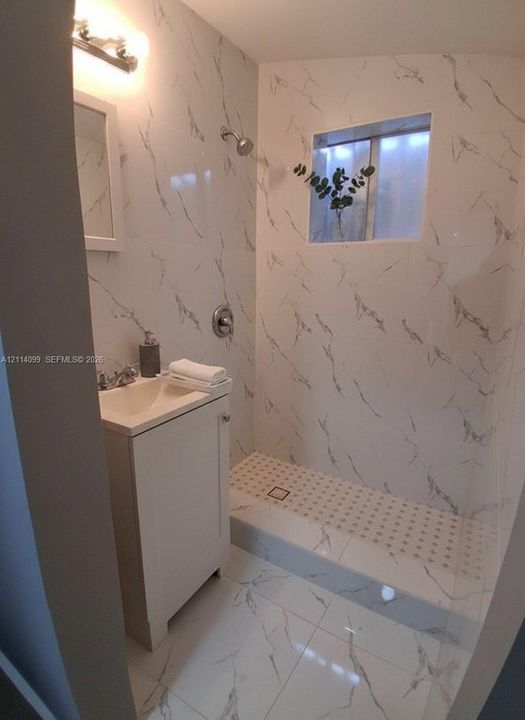 Full-height wall tiling with a veined pattern, walk-in shower with a low-profile threshold, high-gloss floor tiling, vanity cabinet with a integrated sink, and wall-mounted vanity lighting