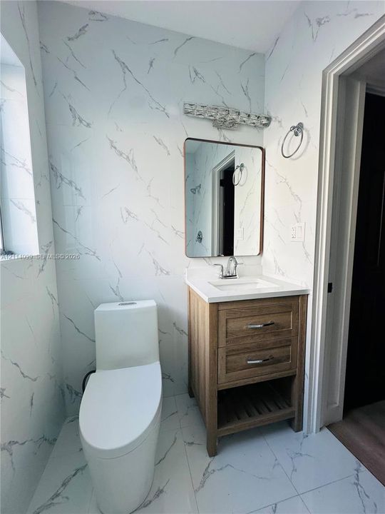 Full-height stone-patterned wall tile, wood-finish vanity with two drawers and lower open shelving, integrated white countertop with rectangular sink, polished chrome faucet, and rectangular mirror