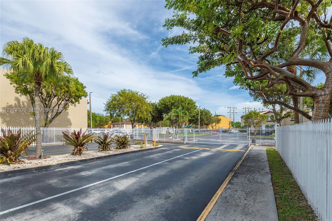 Gated entryway featuring a paved access road, white perimeter fencing, and mature landscaping