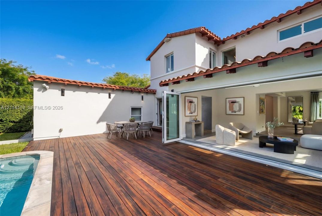 Spanish-style exterior featuring a terra cotta tile roof, expansive wood deck, swimming pool with stone coping, and large-format bi-fold glass doors