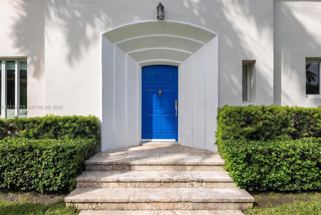 Stucco exterior entrance featuring an arched recessed doorway, paneled front door, stone steps, and manicured shrubbery