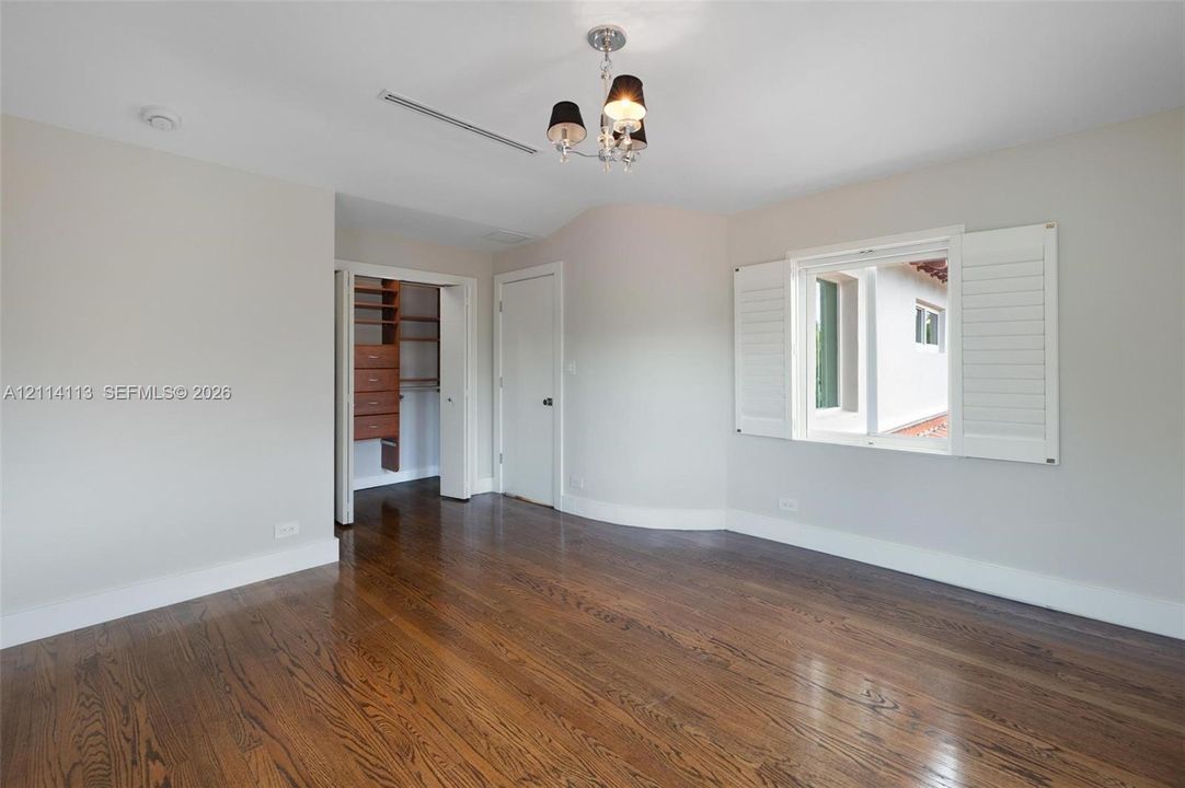 Room featuring wood-finish flooring, white plantation shutters, a multi-light chandelier, and a walk-in closet with built-in wooden shelving and drawers