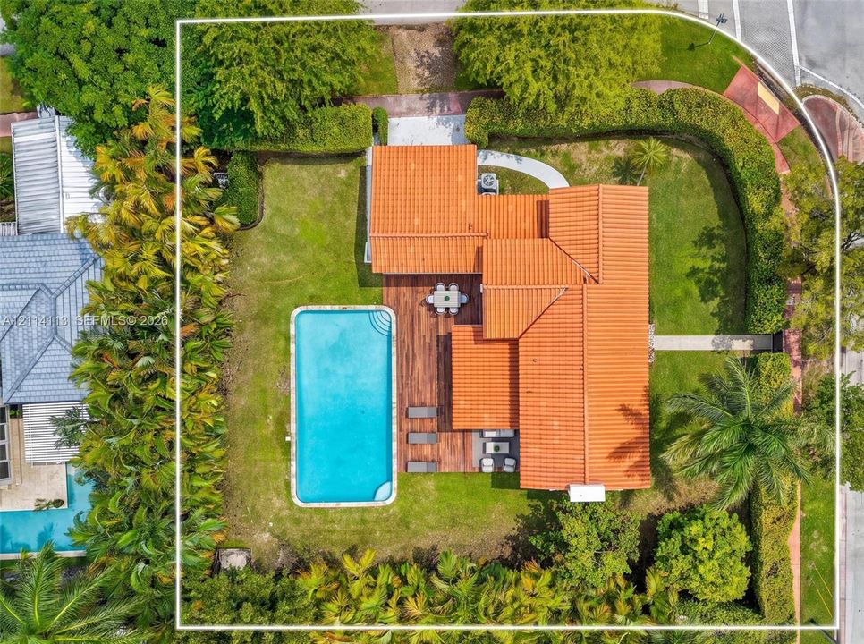 Aerial perspective of a corner lot property featuring a barrel tile roof, an in-ground swimming pool, wood-finish decking, and perimeter privacy hedging