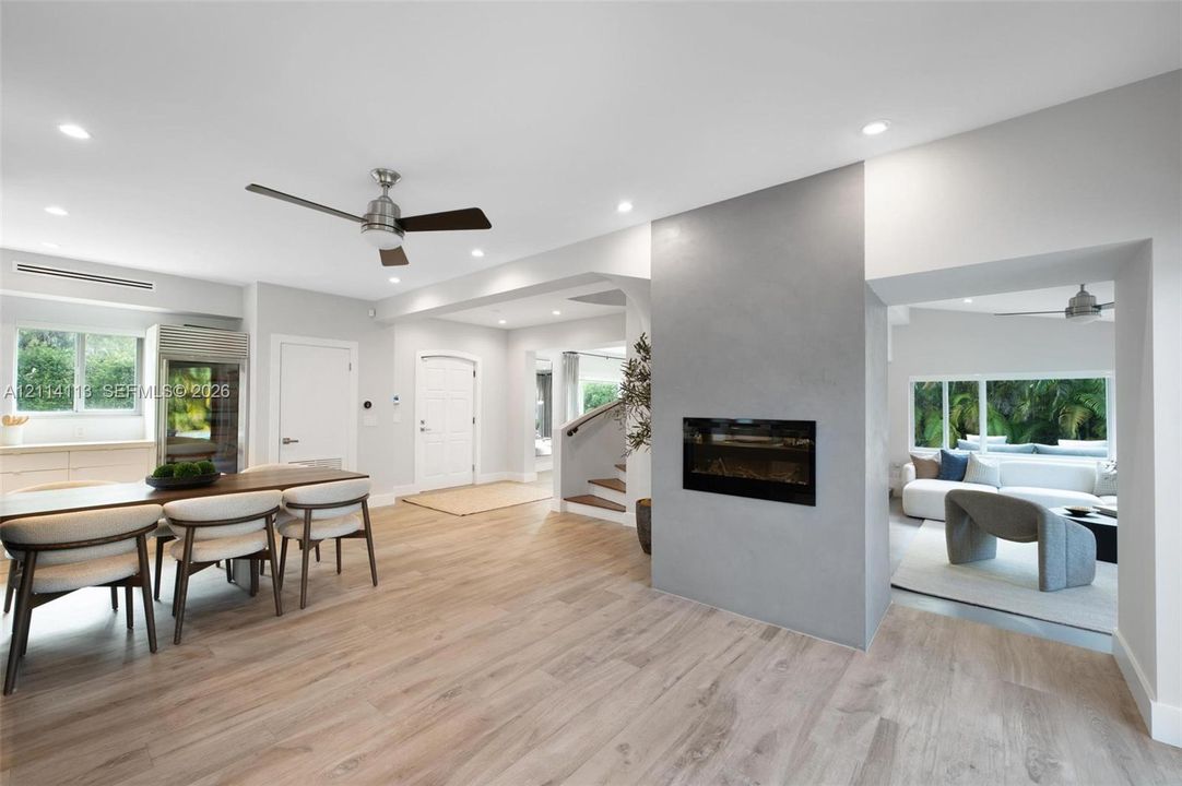 Light-toned wood-finish flooring, central fireplace with a grey smooth-finish surround, recessed ceiling lighting, ceiling fans, and multi-paned windows