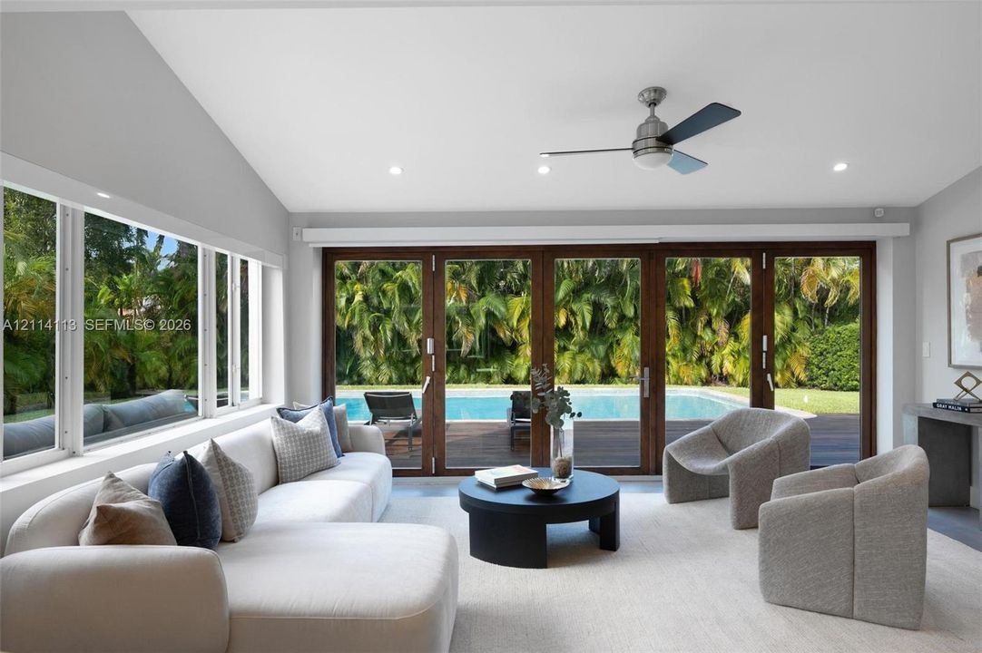 Vaulted ceilings featuring recessed lighting, a modern ceiling fan, expansive multi-panel sliding glass doors, and large windows encompassing exterior pool and landscape elements