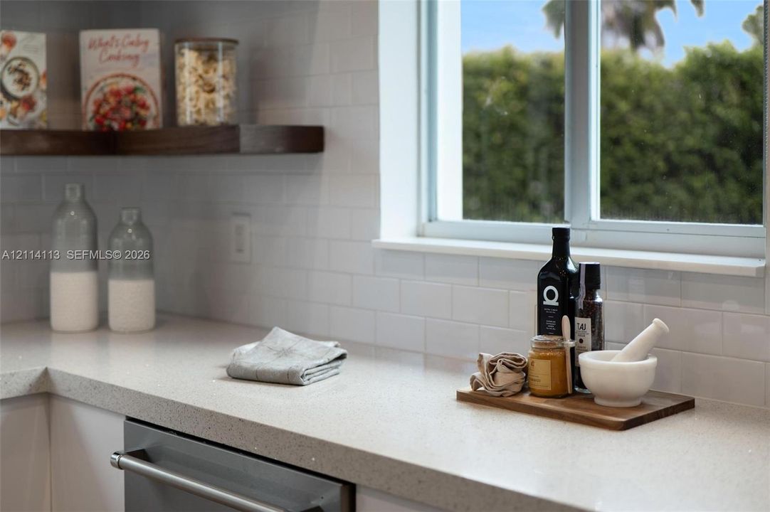 Kitchen featuring light-toned solid surface countertops, white subway tile backsplash, stainless steel dishwasher, and floating wood-finish shelving