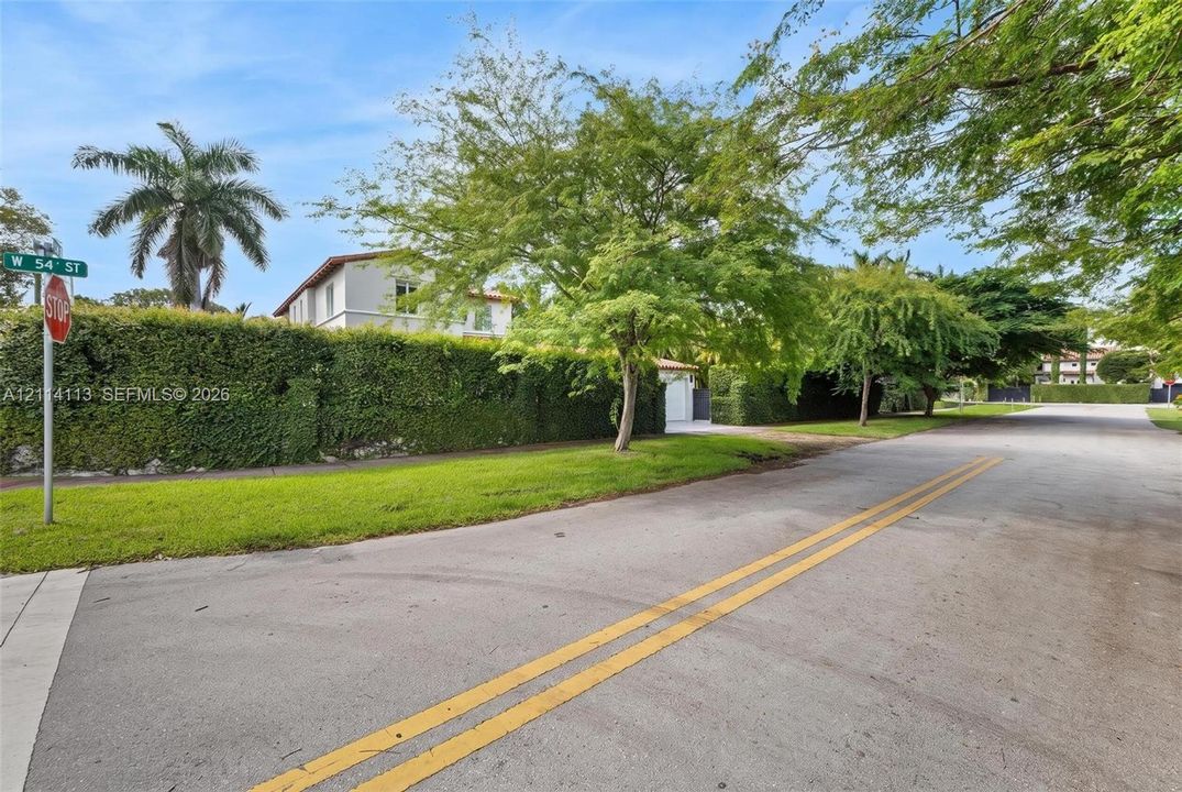 View of the side W 54th street featuring a tall perimeter privacy hedge, manicured lawn, and mature trees