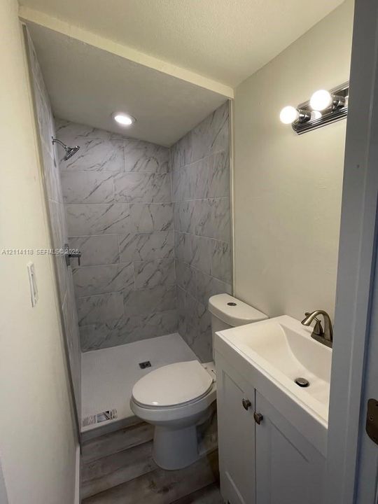 Bathroom featuring a shower stall with stone-patterned wall tile, white vanity cabinet with a integrated sink, polished metal hardware, wood-finish flooring, and wall-mounted vanity lighting