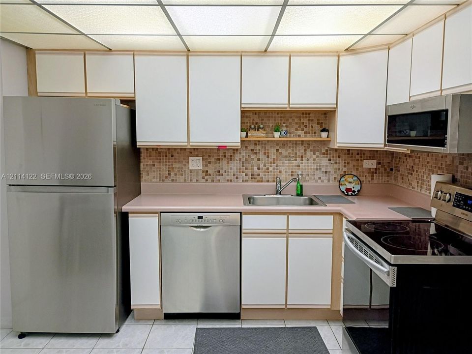 Kitchen featuring stainless steel appliances, flat-panel cabinetry, mosaic tile backsplash, solid surface countertops, and a grid-panel drop ceiling