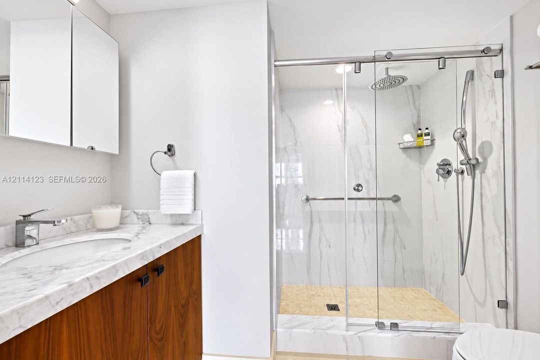 Bathroom featuring a wood-finish vanity with a stone-top integrated sink, stone-surround shower with a glass sliding door, rainfall showerhead, and a mirrored medicine cabinet