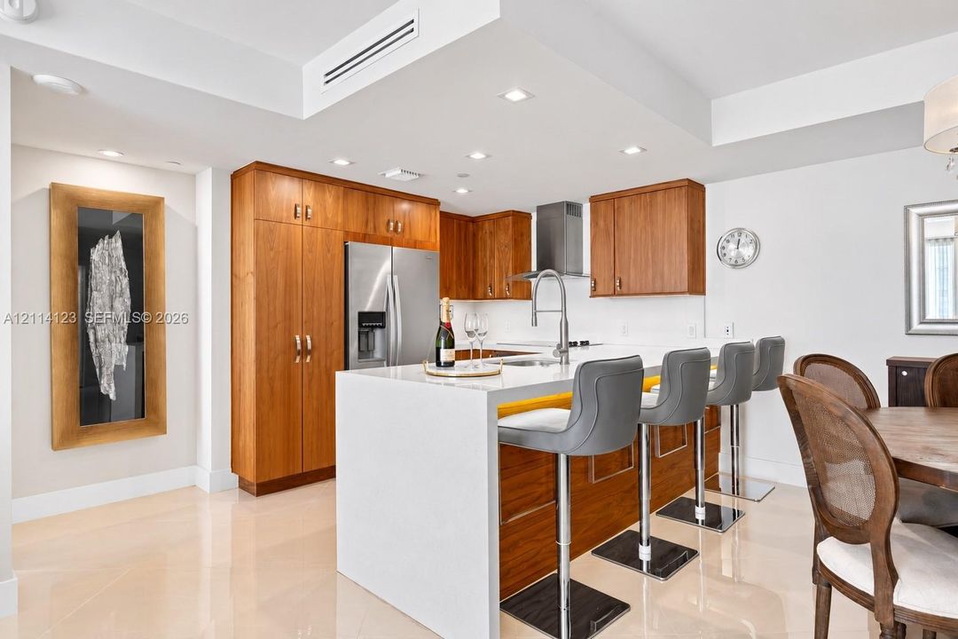 Kitchen featuring custom wood cabinetry, an expansive waterfall-edge island with stone-surface countertop, stainless steel appliances, recessed ceiling lighting, and polished stone flooring