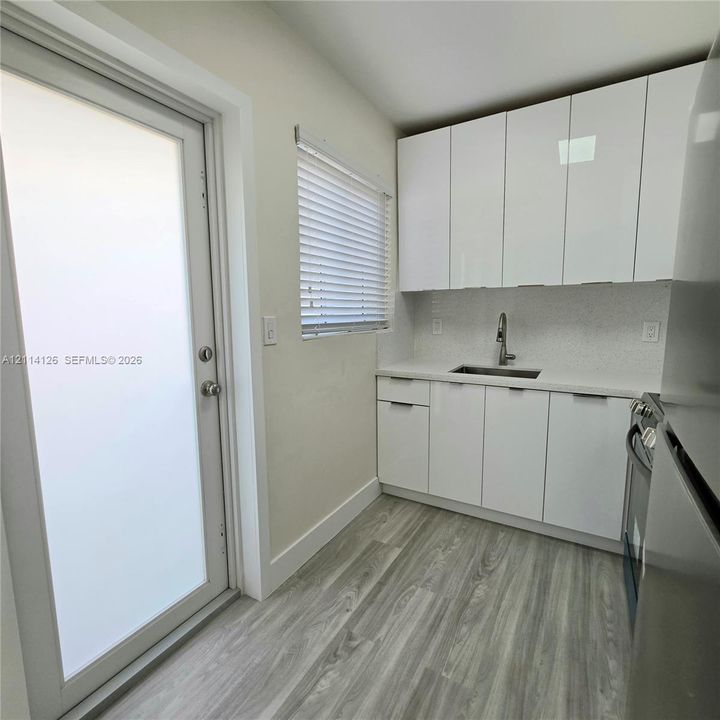 Kitchenette featuring high-gloss white flat-panel cabinetry, light-toned stone-finish countertops, stainless steel undermount sink with a gooseneck faucet, wood-finish flooring, and a frosted glass exterior door
