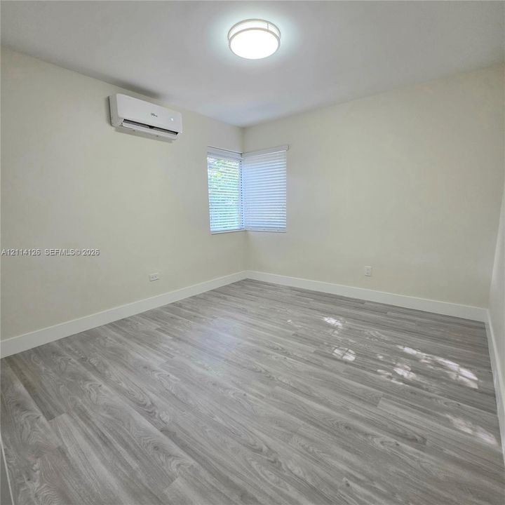 Neutral-toned walls, wood-finish flooring, wall-mounted split system air conditioner, corner window with horizontal blinds, and flush-mount ceiling lighting