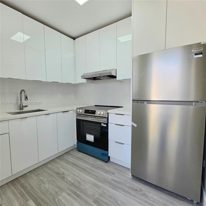 Modern kitchen featuring high-gloss white cabinetry, white stone-finish countertops, stainless steel appliances, and wood-finish flooring