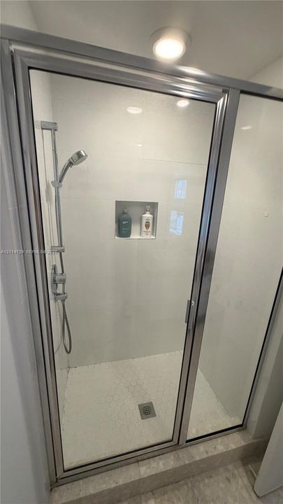 Walk-in shower featuring a chrome hand-held showerhead, glass enclosure, built-in wall niche, white tiled walls, and textured tile flooring
