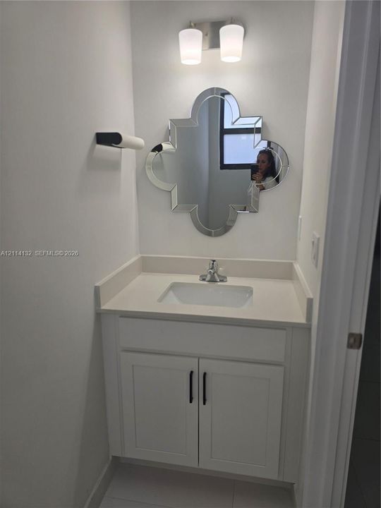Guest 1/2 Bathroom- Vanity featuring white shaker-style cabinetry, matte black hardware, a light-toned countertop with an integrated rectangular sink, a dual-light wall sconce, and a quatrefoil-shaped decorative mirror