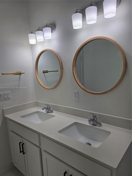 Dual-sink vanity featuring a light-toned solid surface countertop, white shaker-style cabinetry with matte black hardware, circular wood-framed mirrors, and dual three-light wall fixtures with frosted glass shades