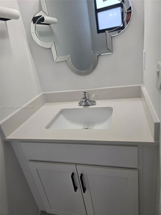 Guest 1/2 Bathroom- Vanity featuring a solid-surface countertop, integrated rectangular sink, white shaker-style cabinetry with matte black hardware, and an ornate-shaped wall mirror