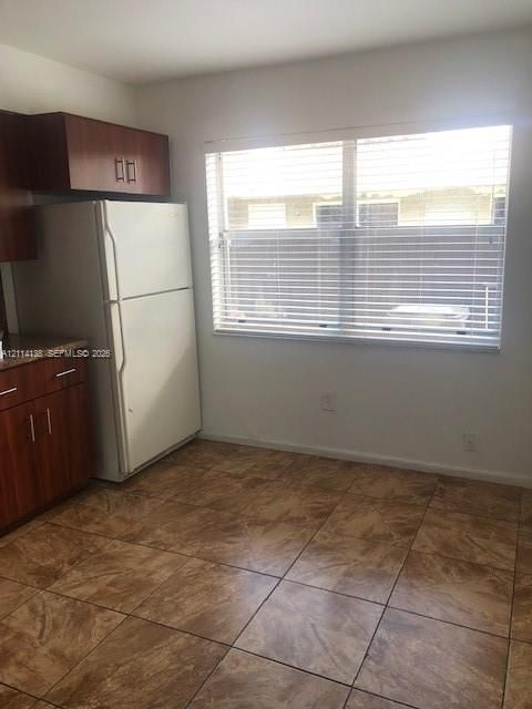 Kitchen featuring wood-finish cabinetry, stone-texture laminate countertops, large-format tile flooring, and a window with horizontal blinds