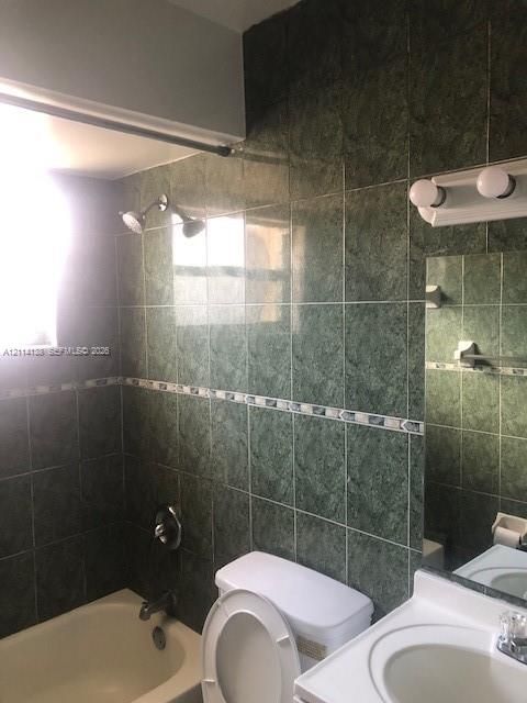Bathroom featuring full-height green ceramic wall tiling with a decorative mosaic border, a built-in bathtub, a white vanity with an integrated sink, and a dual-bulb vanity light fixture