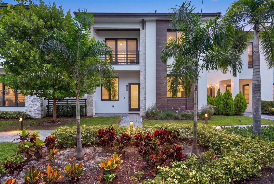 Contemporary two-story exterior featuring smooth stucco siding, stone veneer accents, wood-finish paneling, an upper-level balcony with metal railing, and a paver walkway