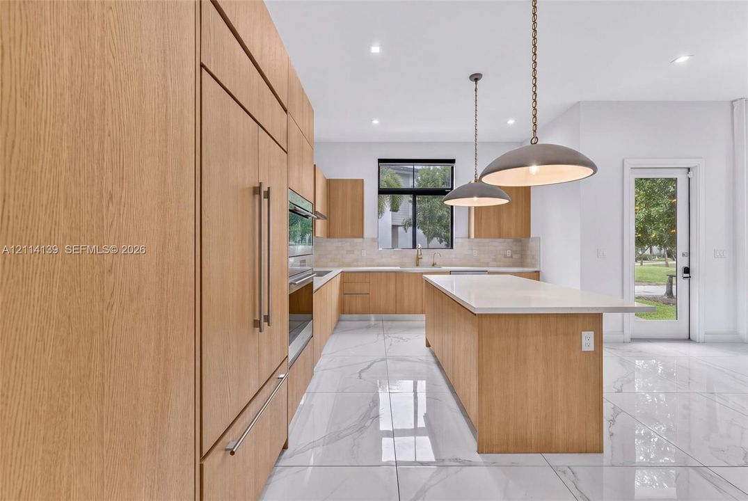 Kitchen featuring flat-panel wood-finish cabinetry, a center island with a stone-surface countertop, built-in panel-ready refrigeration, large-format tile flooring, and dual pendant lighting fixtures