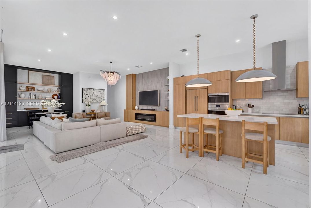 Large-format tile flooring throughout featuring a neutral stone-pattern, light wood cabinetry, integrated stainless steel appliances, decorative pendant lighting over the kitchen island, and a floor-to-ceiling fireplace surround with built-in millwork