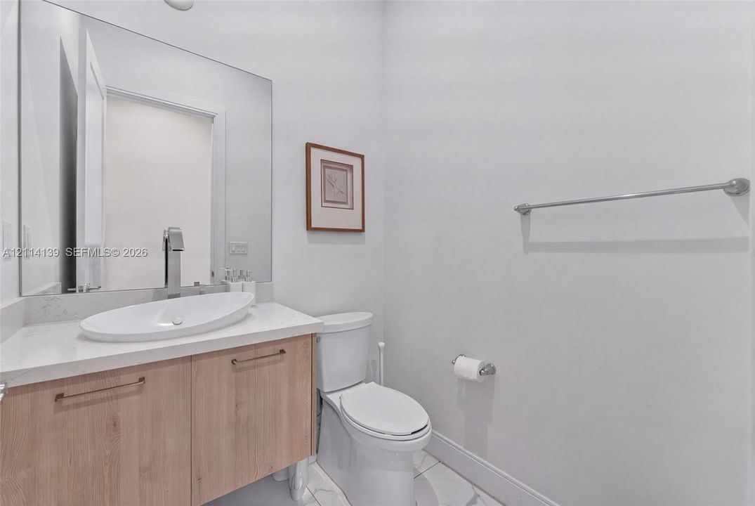 Wood-finish vanity cabinet, white oval vessel sink, light-toned countertop, large frameless mirror, and dual-flush toilet