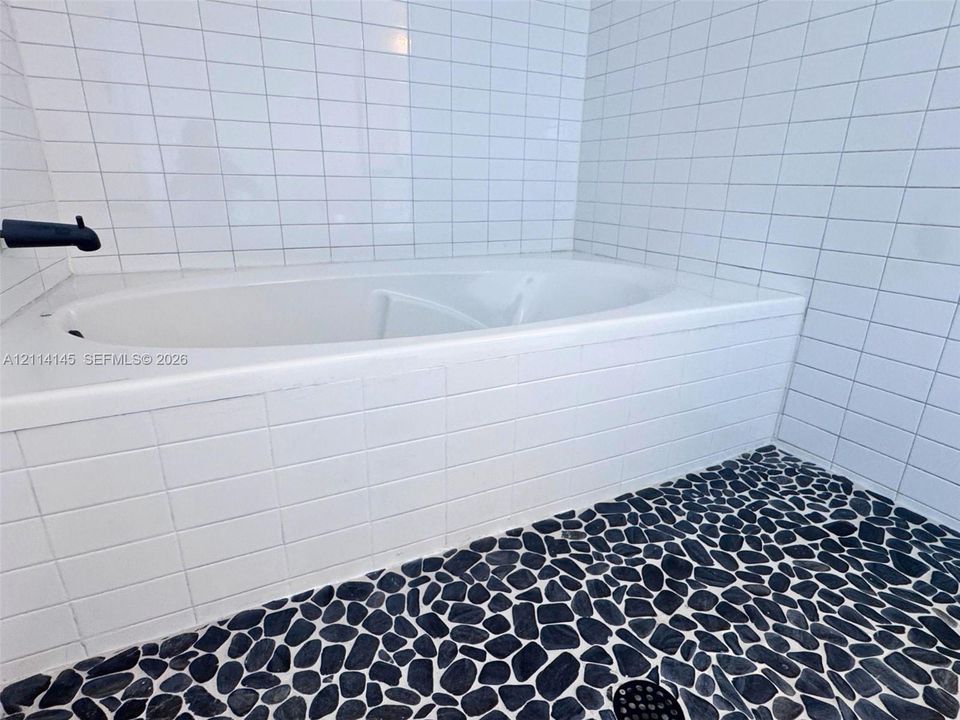 Built-in soaking tub with white square tile surround, matte black wall-mounted faucet, and natural stone pebble flooring