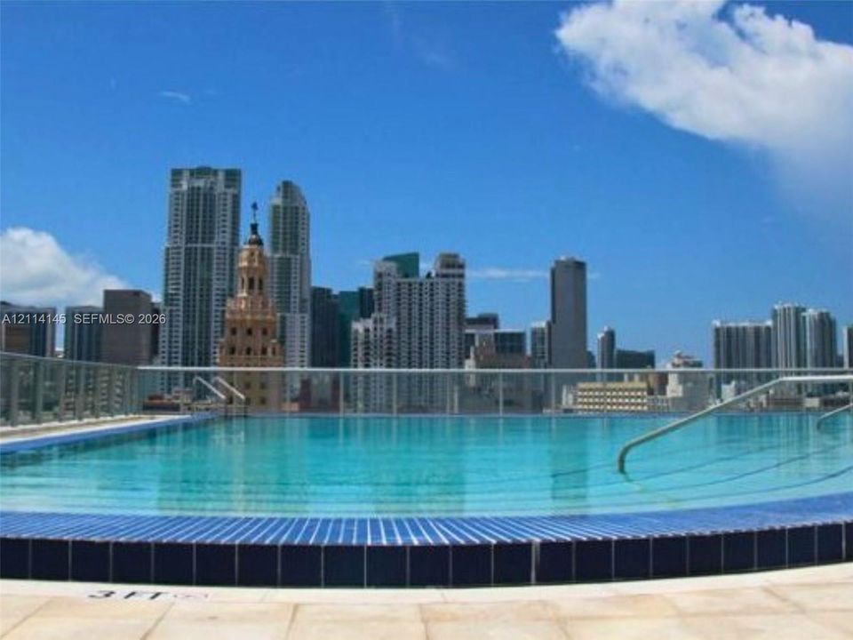 Rooftop swimming pool featuring tiled coping, a perimeter glass railing, and metal handrails, set against an urban skyline