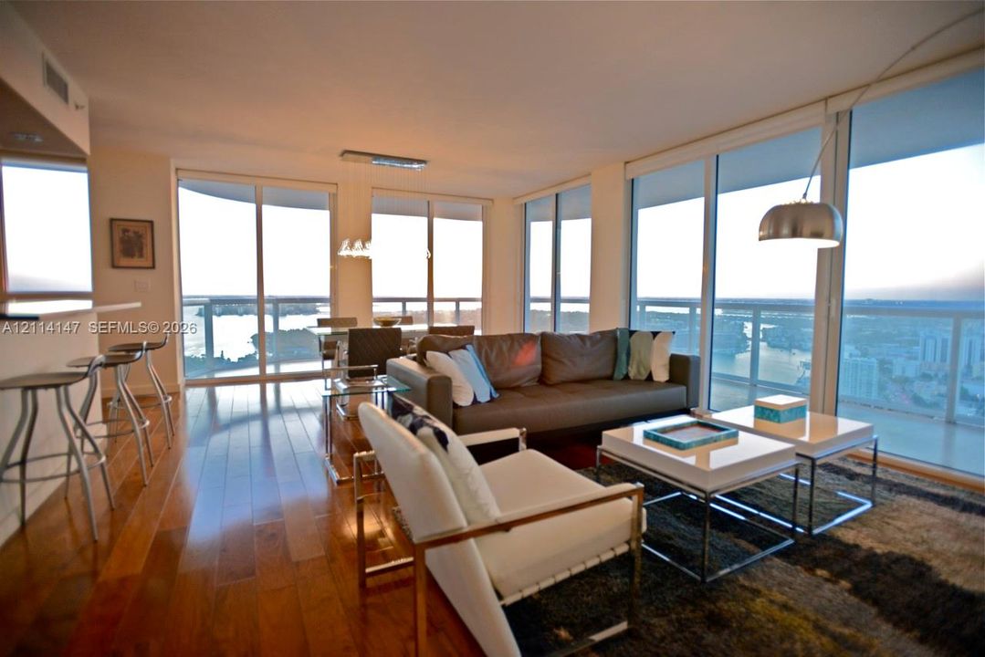 Open-concept interior featuring floor-to-ceiling glass walls, expansive water scenery, wood-finish flooring, and a built-in kitchen breakfast bar