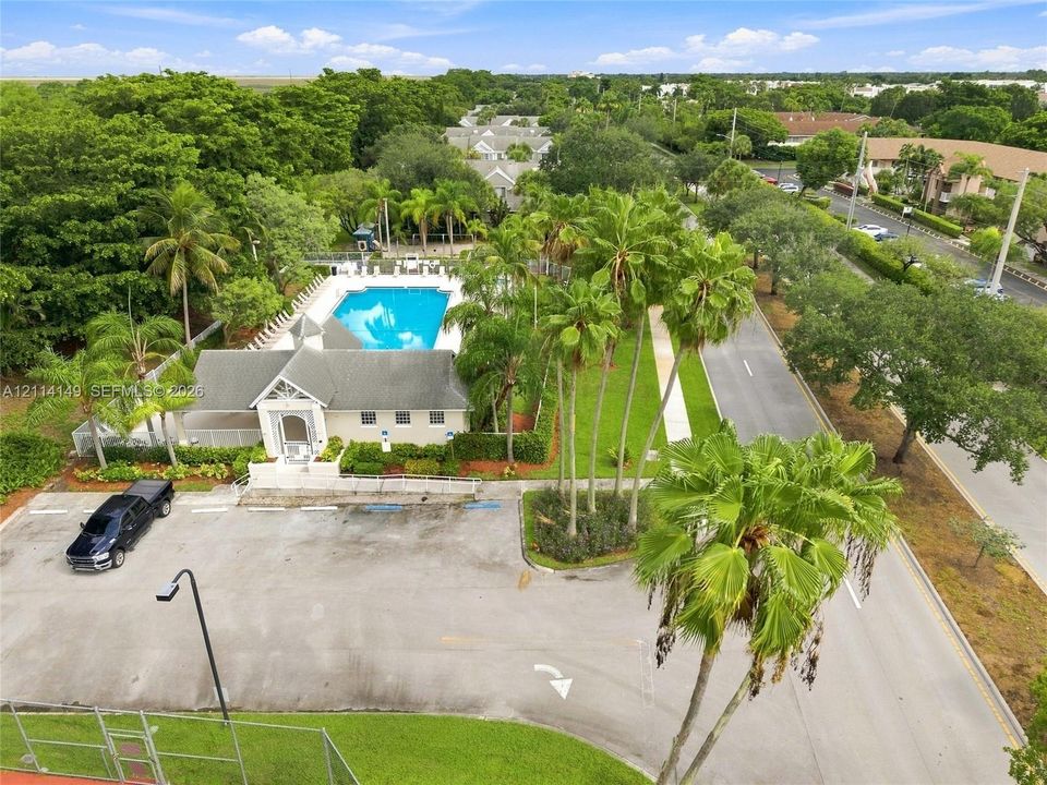 Community recreation center featuring an outdoor swimming pool, paved parking area, mature palm trees, and fenced court